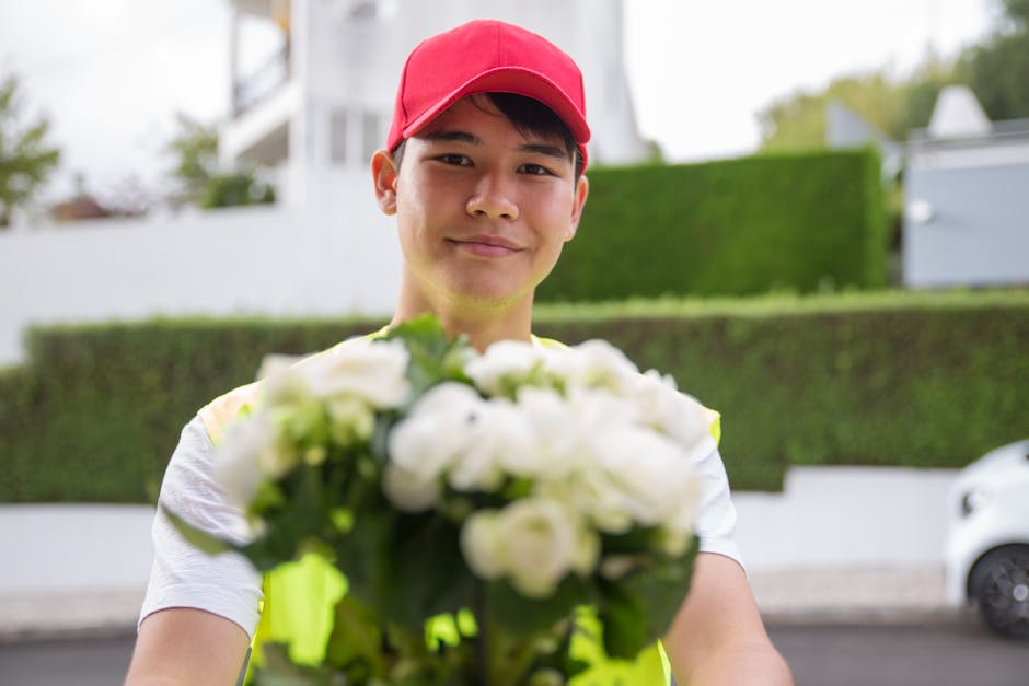 A smiling young delivery person wearing a red cap and a high-visibility jacket is holding a bouquet of fresh white flowers, possibly roses or similar blooms, wrapped in clear plastic. The bouquet is p