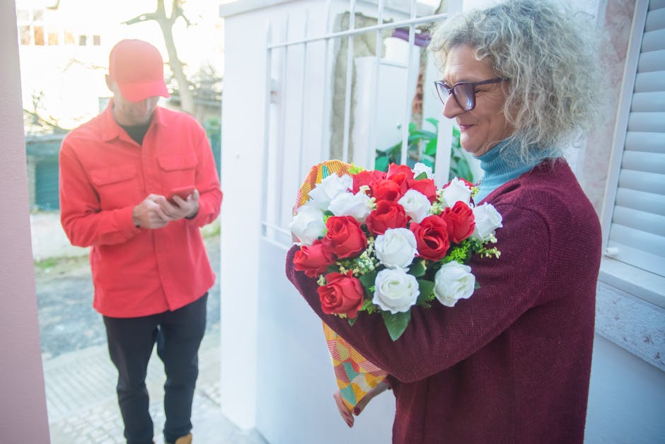 A woman with curly gray hair and glasses is holding a large bouquet of fresh flowers featuring red and white roses, wrapped in colorful patterned paper. She is standing outdoors on a porch near a whit