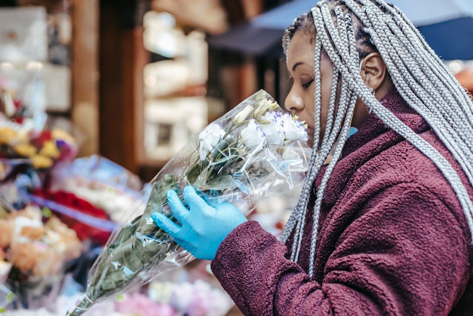 A woman with braided silver hair, wearing a burgundy fleece jacket and blue gloves, is holding a bouquet of white roses wrapped in clear cellophane. She is smelling the flowers in a flower market, wit