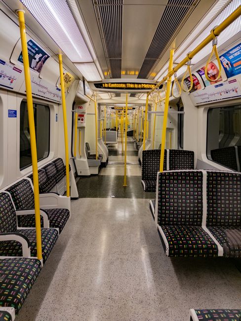 Inside a clean, empty London Underground train car with black and multicolored dotted fabric seats arranged along the sides and facing each other. Bright yellow handrails run along the ceiling and ver