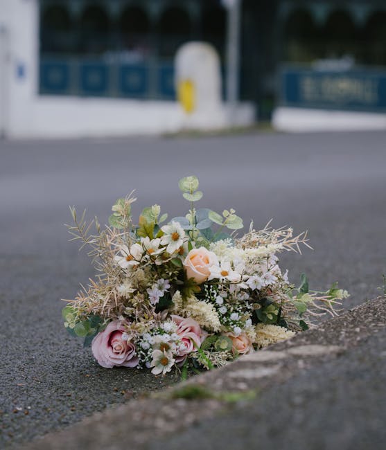 A delicate floral bouquet resting on the pavement near a curb, featuring soft pink roses, small white daisies, and cream-colored filler flowers arranged with eucalyptus and wispy foliage. The arrangem