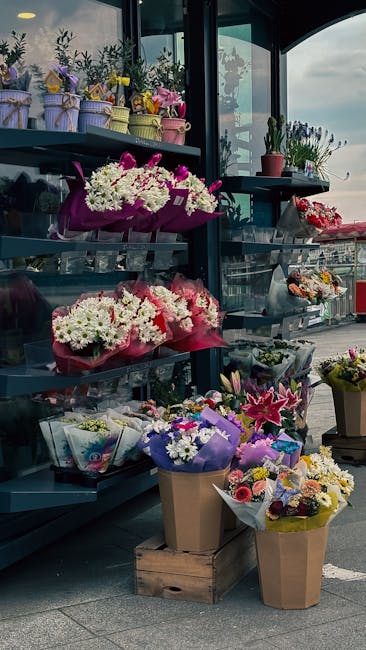 A display of fresh flower bouquets arranged on dark-colored shelving inside a flower shop with large windows, allowing natural light to highlight the vibrant colors. The arrangements include white dai