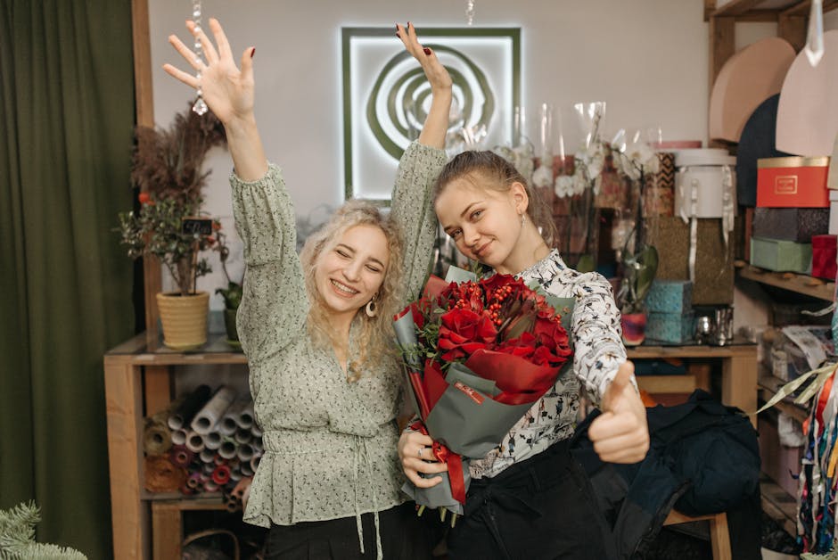 Two young women inside a floral shop are celebrating with a fresh flower bouquet. The woman on the right holds a large, vibrant arrangement of red roses wrapped in grey and red paper, tied with a red 